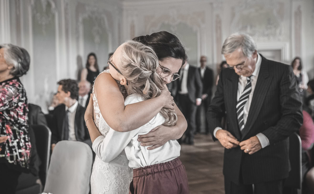 Zwei Personen umarmen sich in einem eleganten Raum mit mehreren sitzenden und stehenden Personen im Hintergrund.