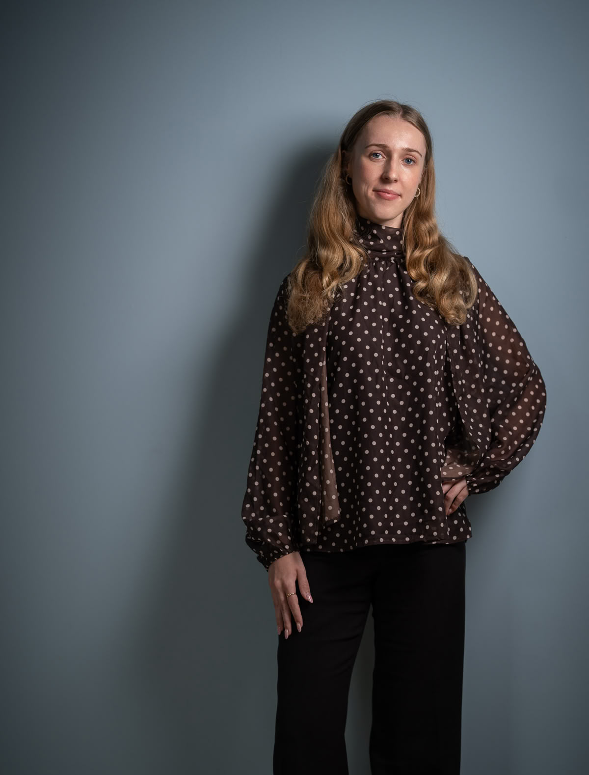 Frau mit langen Haaren trägt eine gemusterte Bluse und schwarze Hose, steht vor einer grauen Wand.