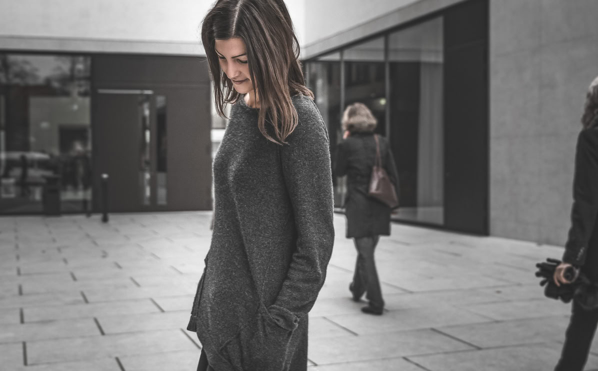 Eine Frau in einem dunklen Pullover steht auf einem Gehweg, während zwei weitere Personen im Hintergrund gehen.