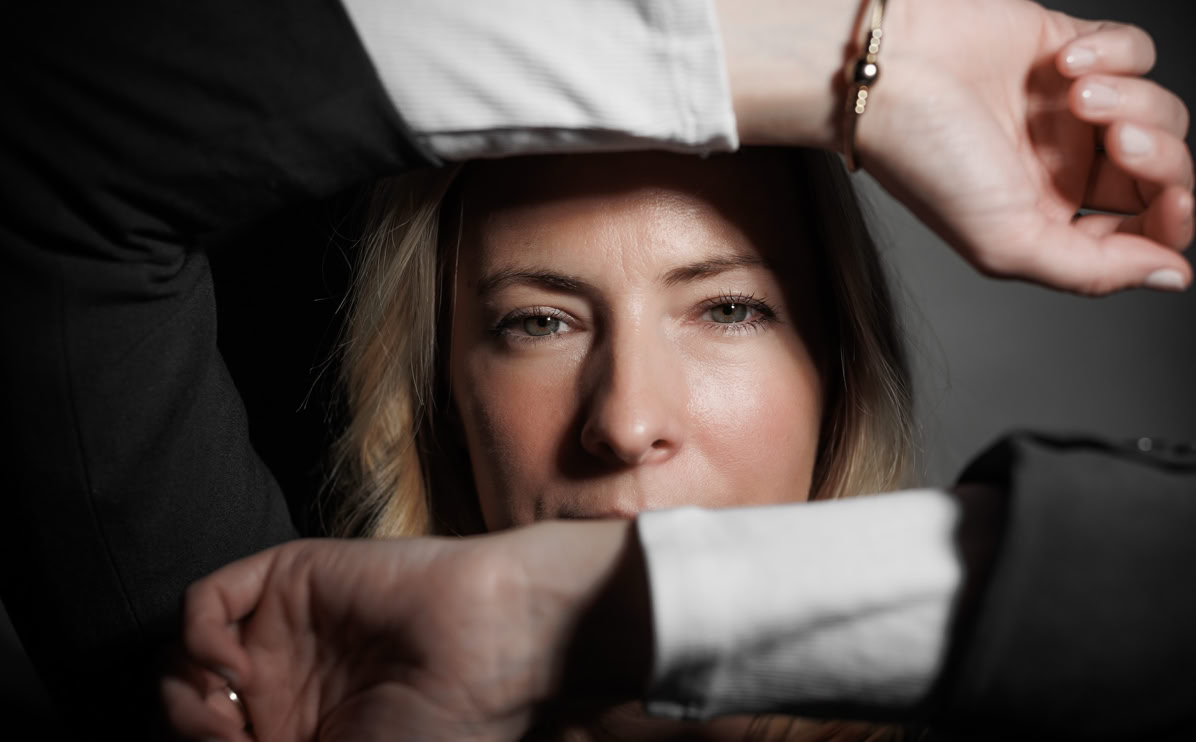 Frau mit blonden Haaren, die durch verschränkte Arme blickt, trägt ein Armband.