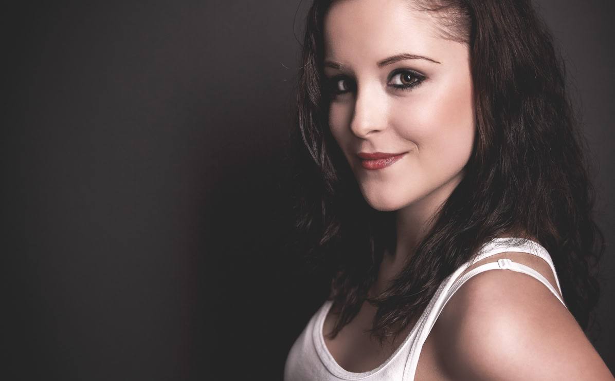 Eine Frau mit langen, dunklen Haaren und einem weißen Oberteil lächelt in die Kamera vor einem grauen Hintergrund.