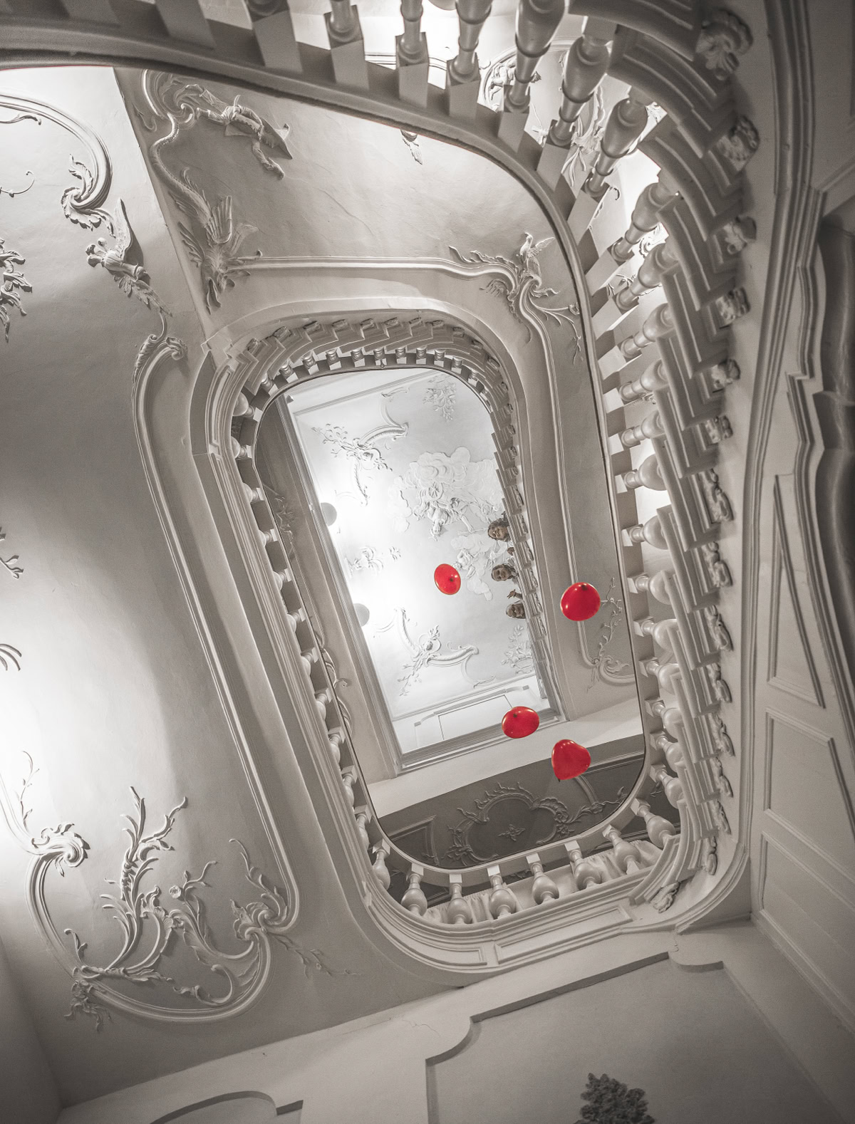 Blick nach oben durch ein spiralförmiges Treppenhaus mit verzierten Wänden und Decken, rote Luftballons schweben in der Mitte.
