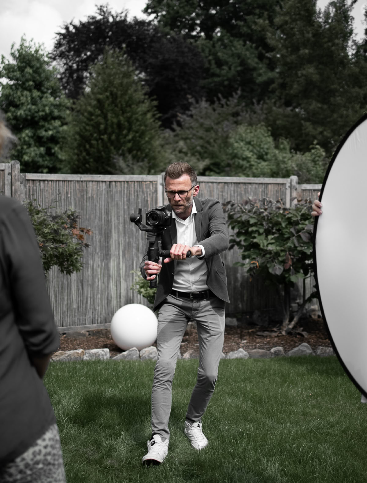 Eine Person mit Kamera fotografiert eine andere Person, die einen weißen Ball hält, während eine dritte Person einen Reflektor hält, im Garten mit Holzzaun und Bäumen im Hintergrund.