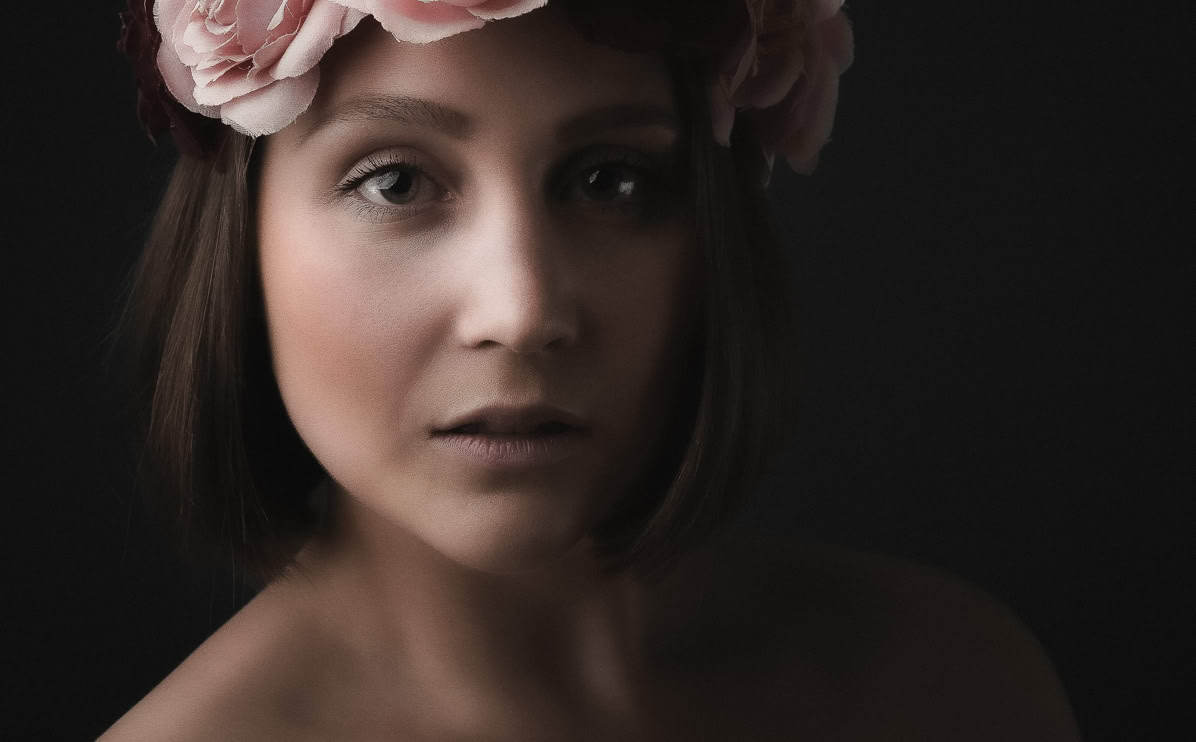 Eine Frau mit kurzen braunen Haaren trägt einen Blumenkranz aus rosa Blüten auf dem Kopf, vor einem dunklen Hintergrund.