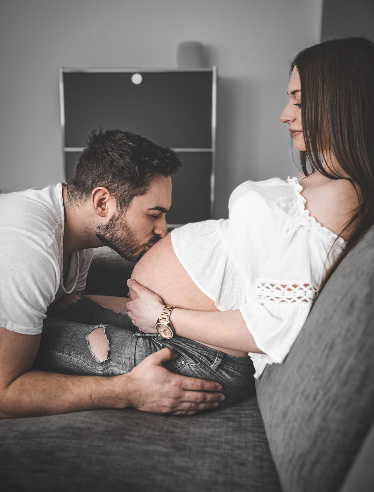 Ein Mann küsst den Bauch einer schwangeren Frau, die auf einem Sofa sitzt.