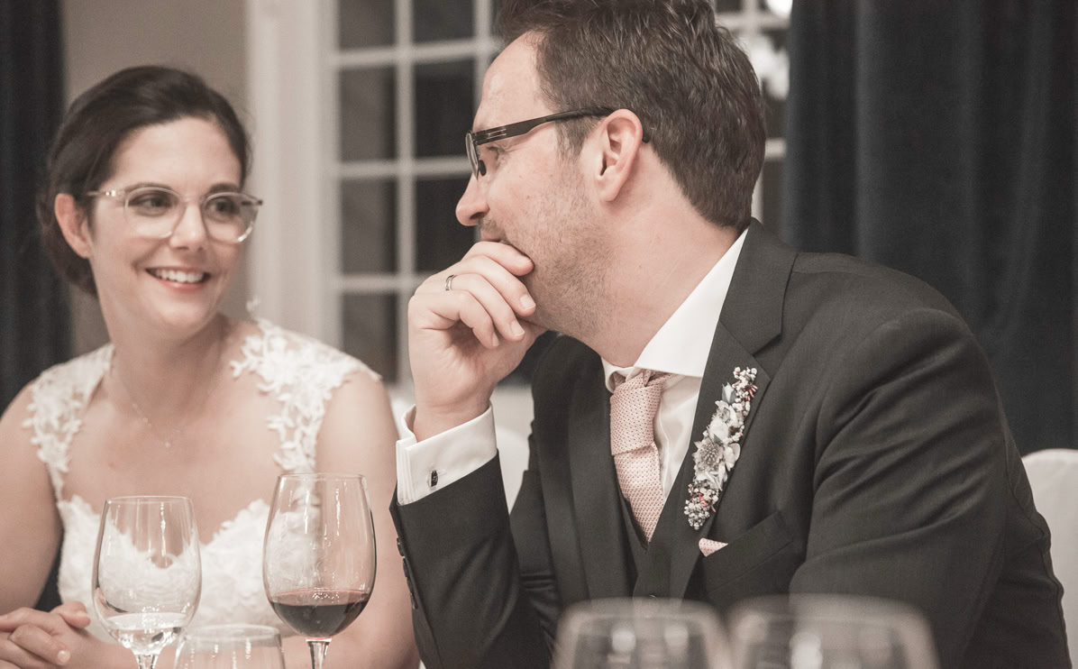 Ein lächelndes Paar sitzt an einem Tisch mit Weingläsern, beide tragen elegante Kleidung.