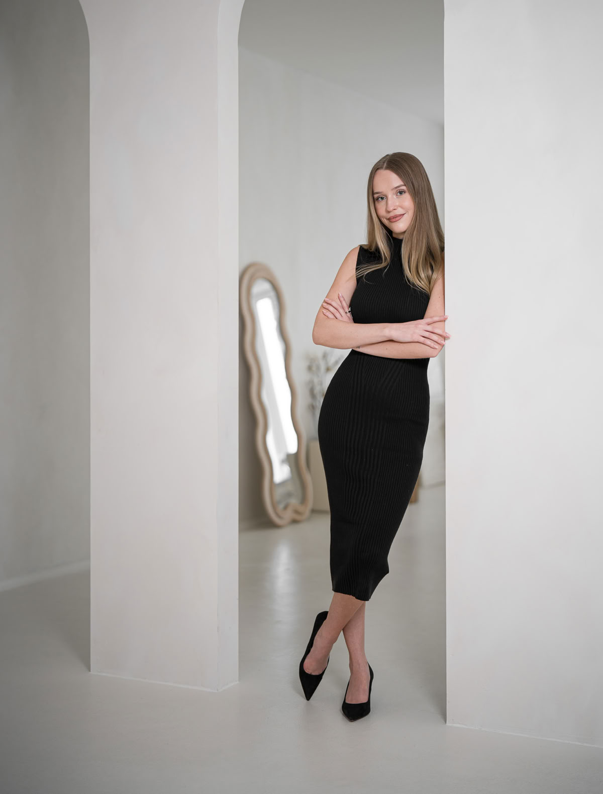 Frau in schwarzem Kleid lehnt an einem weißen Bogen, mit einem großen Spiegel im Hintergrund.