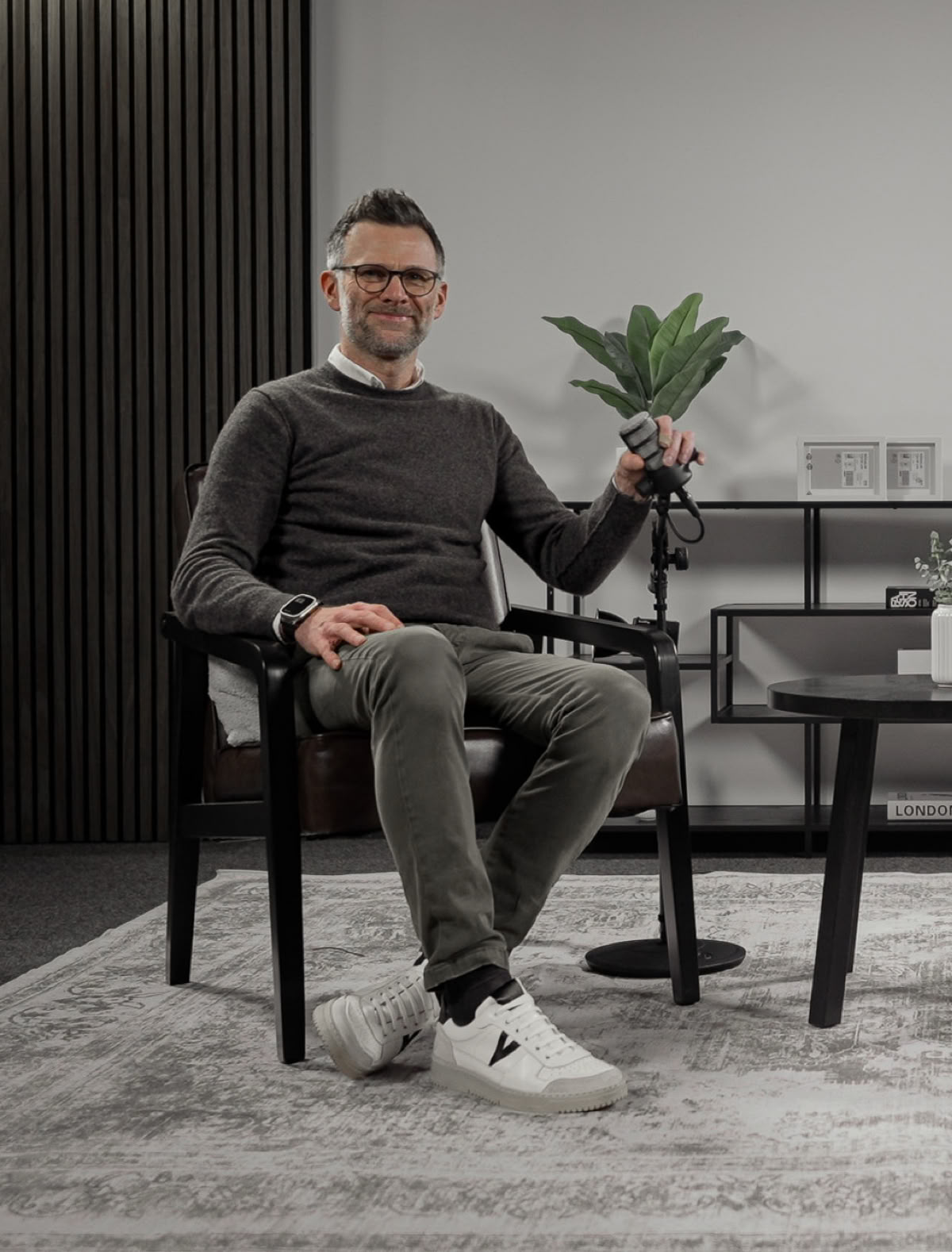 Ein Mann sitzt auf einem Stuhl in einem modern eingerichteten Raum mit einem Tisch, einem Mikrofon und Pflanzen im Hintergrund.