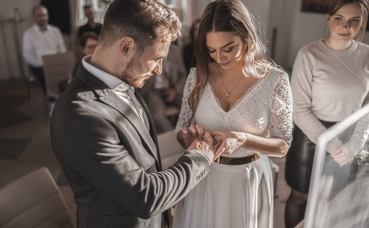 Ein Mann im Anzug steckt einer Frau im weißen Kleid einen Ring an den Finger, während mehrere Personen im Hintergrund zuschauen.