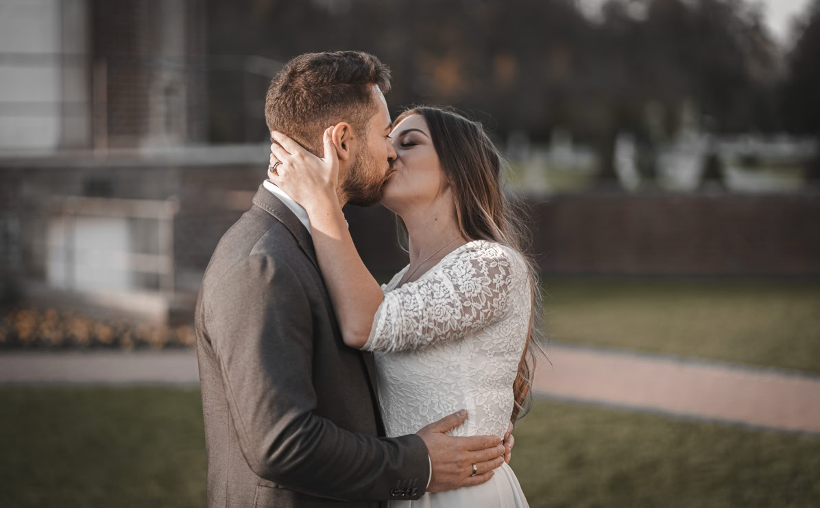 Ein Mann und eine Frau küssen sich im Freien, die Frau trägt ein weißes Kleid, der Mann einen dunklen Anzug.