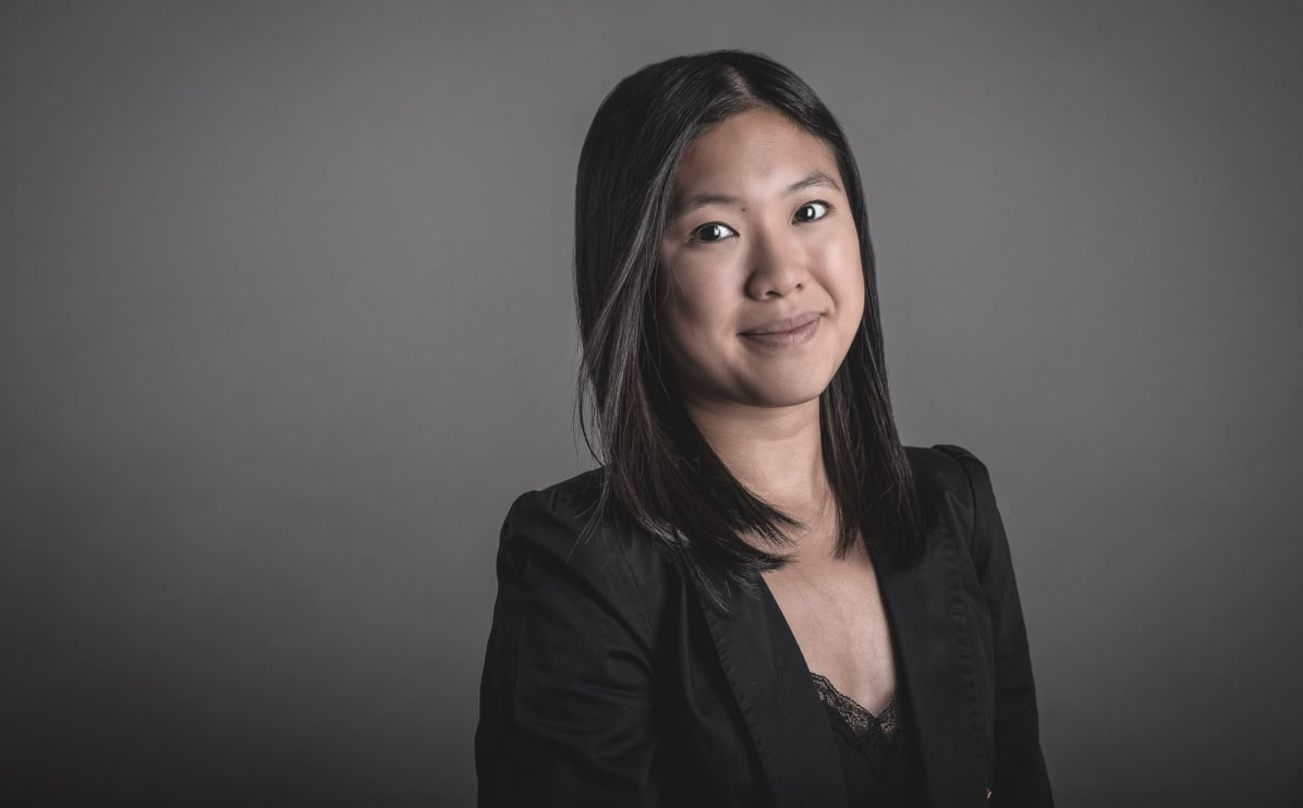 Eine Frau mit langen, dunklen Haaren trägt einen schwarzen Blazer und lächelt vor einem grauen Hintergrund.