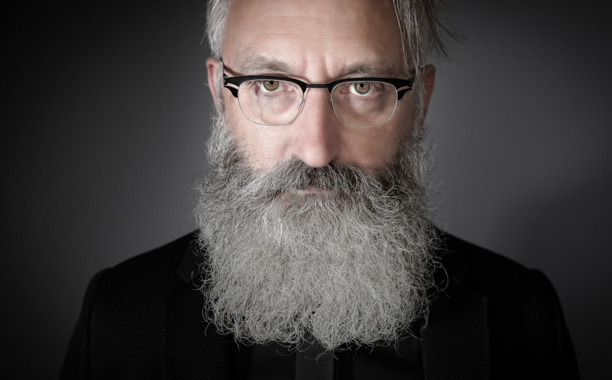 Ein Mann mit grauem Bart und Brille vor grauem Hintergrund.