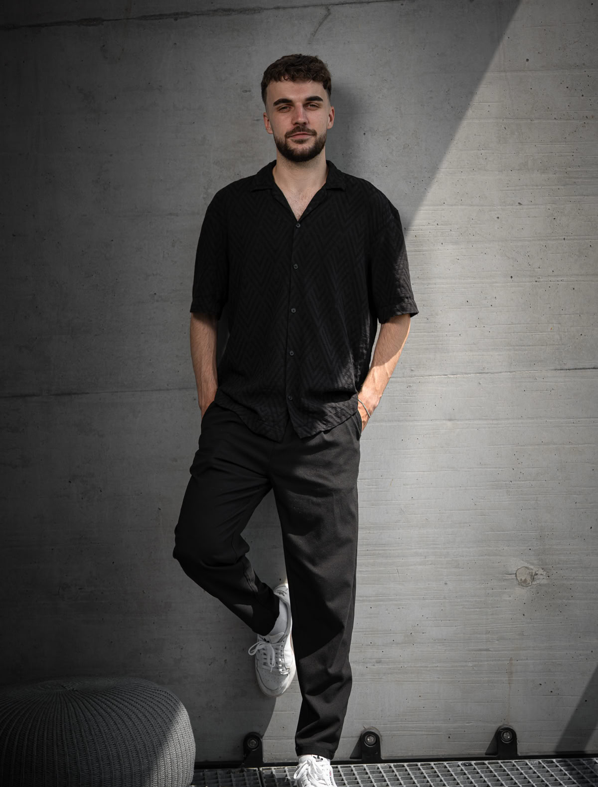 Mann in schwarzer Kleidung und weißen Schuhen lehnt an einer grauen Betonwand.