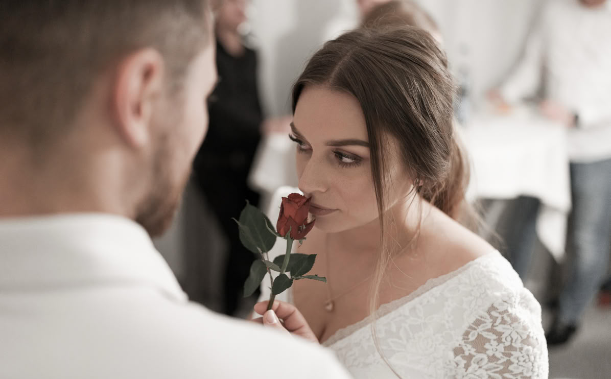Frau in weißem Kleid riecht an einer roten Rose, während sie einen Mann ansieht; unscharfer Hintergrund mit mehreren Personen.
