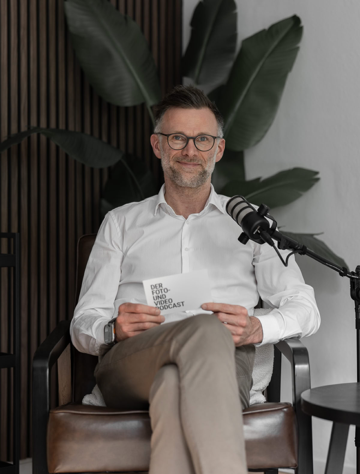 Ein Mann sitzt auf einem Stuhl in einem modernen Raum mit Pflanzen im Hintergrund, hält ein Blatt Papier und hat ein Mikrofon vor sich.