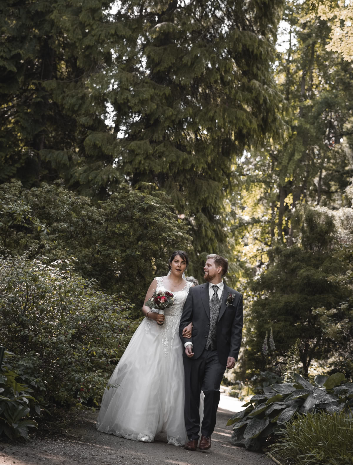 Ein Paar in Hochzeitskleidung geht auf einem Waldweg, umgeben von hohen Bäumen und üppigem Grün.