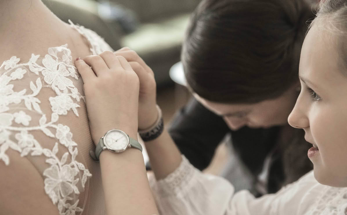 Personen helfen einer Frau, ein weißes Spitzenkleid zu schließen.