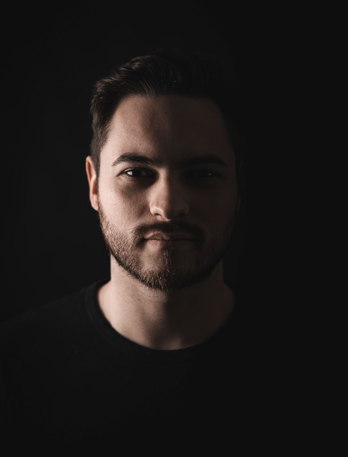 Porträt eines Mannes mit Bart, der in einem dunklen Raum steht, mit Licht auf seinem Gesicht.